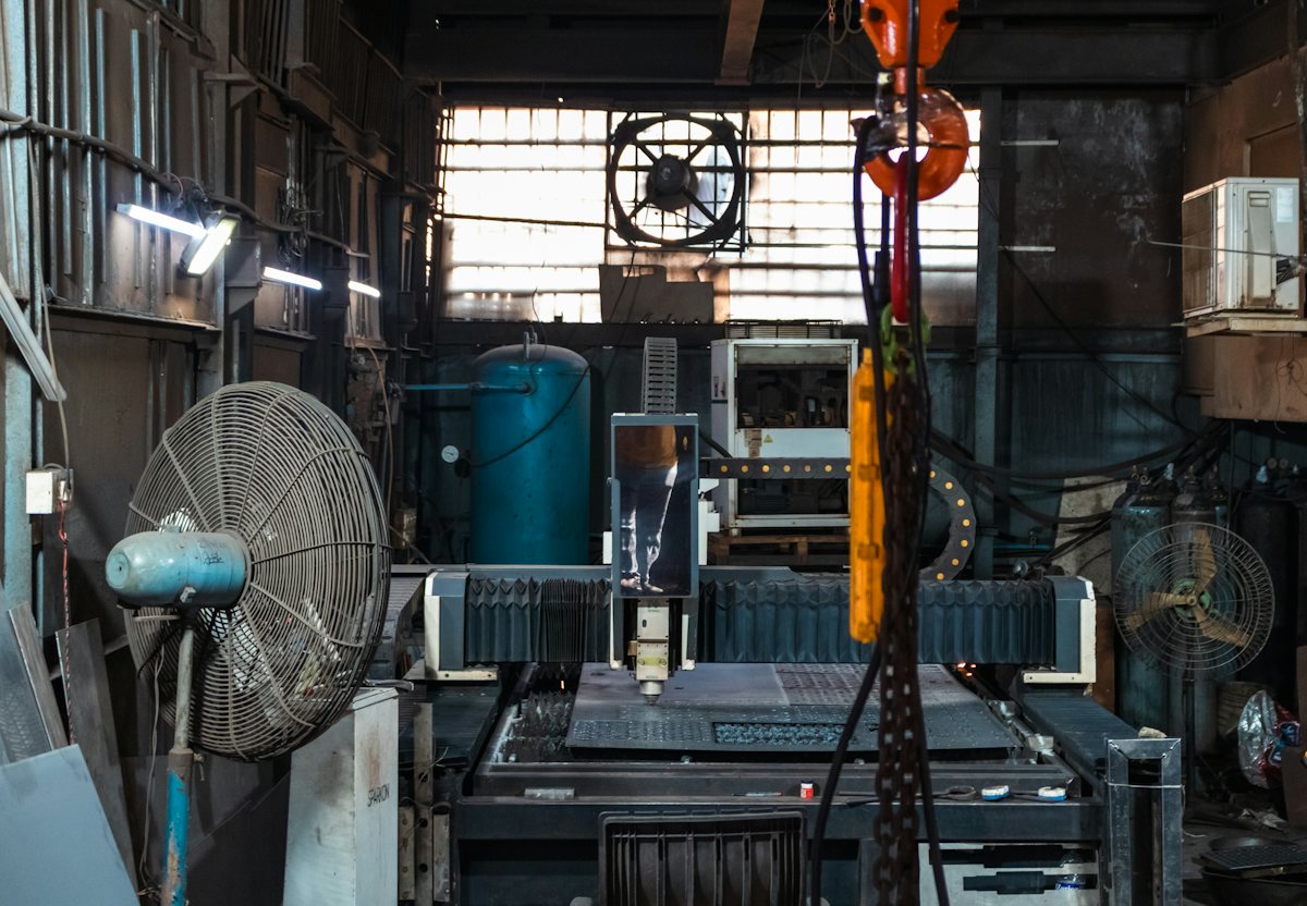 A small machine shop: workbenches and machines under tall factory windows, a big fan on the wall